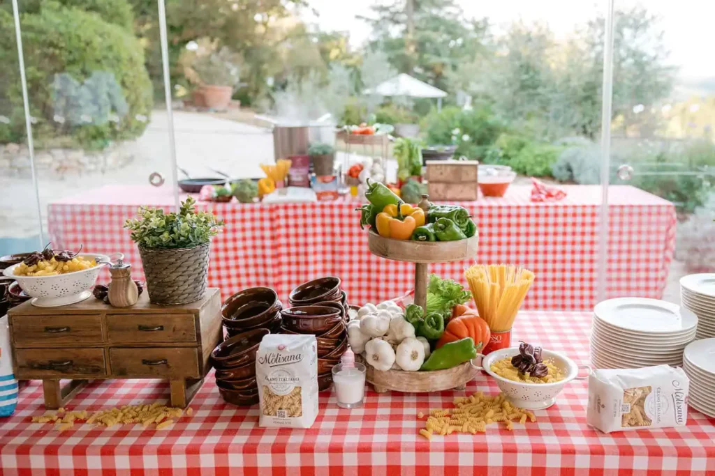 Table set with rustic-chic style red and white checked tablecloth Agnesi pasta, white colander, fresh vegetables (tomatoes, peppers), garlic, a wooden pepper grinder, and an aromatic plant. The setting suggests a traditional Italian cuisine with show-cooking decorations.
