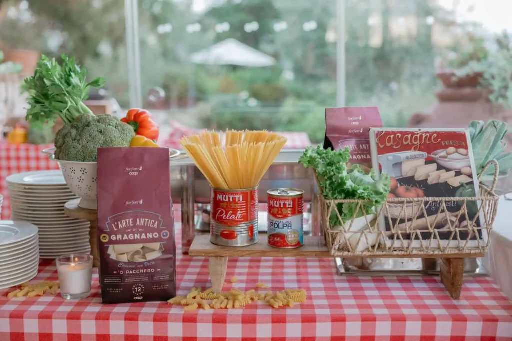 Tavolo apparecchiato con tovaglia a quadretti bianchi e rossi in stile rustic-chic
Pasta Agnesi, colapasta bianco, verdure fresche (pomodori, peperoni), aglio, un macinino per pepe in legno e una pianta aromatica.
L'ambiente suggerisce una cucina tradizionale italiana con Decorazioni per show-cooking.